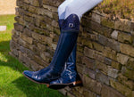 A navy patent De Niro riding boot with a decorated top, worn against a dry-stone wall
